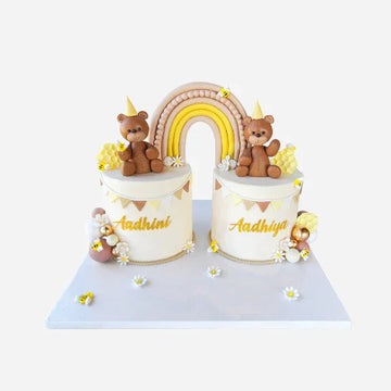 Two small cylindrical cakes with teddy bears and a rainbow on top, on a white background.